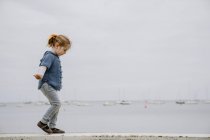 Вид збоку маленької дівчинки з простягнутими руками, що йдуть на кордоні з спокійним морем і сірим небом — стокове фото