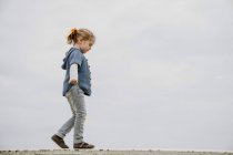 Vista lateral da menina com braços estendidos andando na fronteira contra o mar calmo e céu cinza — Fotografia de Stock