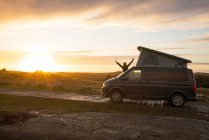 Женский силуэт с открытыми руками, стоящими на трейлере каравана на одинокой проселочной дороге на закате в Уэльсе — стоковое фото