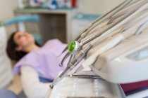 Сучасні інструменти стоматолога, такі як дриль, гострий, скребок і різьбяр з молодою пацієнткою, що лежить на фоні в стоматологічній клініці — стокове фото
