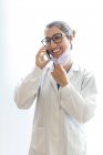 Junge glückliche schöne Zahnärztin in medizinischem Gewand und Brille, die am Telefon spricht und isoliert auf weißem Hintergrund wegschaut — Stockfoto