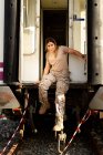 Fuerte soldado femenino mirando a la cámara y sonriendo mientras está sentado en la puerta de un vehículo militar gruñón en el campo - foto de stock