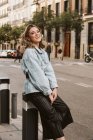 Séduisante femme heureuse en tenue tendance assise sur la borne dans la rue de la ville — Photo de stock