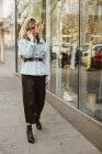 Приваблива молода жінка в стильному вбранні дивиться на одяг за вікном магазину під час прогулянки по міській вулиці — стокове фото