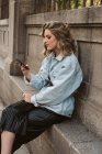 Giovane signora in elegante vestito smartphone di navigazione mentre seduto vicino alla recinzione di pietra del parco della città — Foto stock