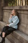 Молода леді в стильному Екіпіровка на мобільному телефоні сидячи біля кам'яного паркану міського парку — стокове фото