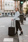 Стильна молода жінка з валізою дивиться геть, перетинаючи місто вулиці — стокове фото