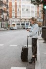 Стильная молодая женщина с помощью мобильного телефона с чемоданом при пересечении улицы города — стоковое фото
