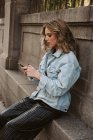 Молодая леди в стильном наряде просматривает смартфон, сидя рядом с каменным забором городского парка — стоковое фото