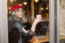 Молодая женщина в красном берете пьет горячий напиток и смотрит в окно, просматривая ноутбук в кафе — стоковое фото