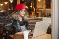 Junge Frau in roter Baskenmütze mit Heißgetränk und Laptop sitzt im Café — Stockfoto