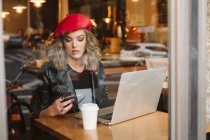 Jeune femme branchée en béret rouge en utilisant un téléphone portable tout en étant assis à table avec ordinateur portable au restaurant — Photo de stock