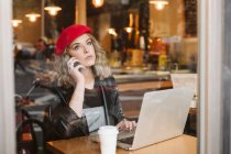 Mujer joven de moda en boina roja en el teléfono mientras está sentado en la mesa con el ordenador portátil en el restaurante - foto de stock