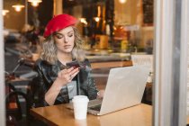Menina na moda em boina vermelha usando telefone celular enquanto sentado à mesa com laptop no restaurante — Fotografia de Stock