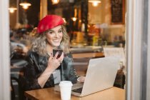 Trendy happy young female in red beret holding mobile phone while sitting at table with laptop in restaurant — Stock Photo