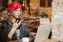 Giovane donna alla moda in berretto rosso utilizzando il telefono cellulare mentre seduto a tavola con computer portatile nel ristorante — Foto stock