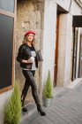 Stylische junge Frau mit roter Baskenmütze hält Imbissgetränk in der Hand, während sie aus dem Café auf der Stadtstraße geht — Stockfoto