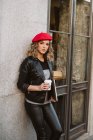 Elegante joven hembra en boina roja sosteniendo una bebida para llevar apoyada en la pared de una cafetería en la calle de la ciudad - foto de stock