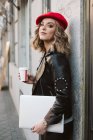 Стильная молодая женщина в красном берете с напитком на вынос, прислонившаяся к стене кофейни на городской улице — стоковое фото
