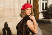 Sensuale giovane donna in berretto alla moda in possesso di giacca di pelle sulla spalla e guardando la fotocamera mentre in piedi vicino al muro edificio nella giornata di sole sulla strada della città — Foto stock