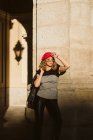 Sensuale giovane donna in berretto alla moda e occhiali tenendo giacca di pelle sulla spalla e guardando la fotocamera mentre in piedi vicino al muro edificio nella giornata di sole sulla strada della città — Foto stock