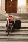 Giovane donna in abito alla moda sorridente mentre seduto sulle scale vicino alla valigia e godendo bevanda da asporto — Foto stock