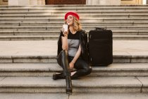 Happy young woman in trendy outfit looking away while sitting on stairs near suitcase and enjoying takeaway beverage — Stock Photo