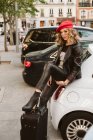 Junge Frau im trendigen Outfit tritt auf Koffer, während sie sich an Auto lehnt — Stockfoto