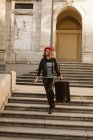 Young woman in trendy outfit with modern sunglasses walking on stairs with suitcase — Stock Photo