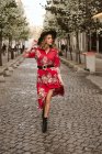 Молодая женщина в стильном платье и шляпе, глядя в сторону во время прогулки по старому тротуару на улице города — стоковое фото