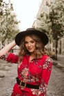 Молода жінка в стильному одязі і капелюсі дивиться на камеру, перебуваючи на старій тротуарі на міській вулиці — стокове фото