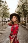 Чувственная молодая женщина в стильном платье и шляпе, глядя в камеру, стоя на старом тротуаре на городской улице — стоковое фото