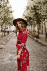 Sensuale giovane donna in abito elegante e cappello guardando la fotocamera mentre in piedi sul marciapiede invecchiato sulla strada della città — Foto stock