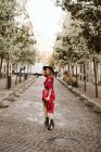 Vue arrière de la jeune femme en robe élégante et chapeau regardant la caméra sur l'épaule tout en marchant sur le trottoir âgé sur la rue de la ville — Photo de stock