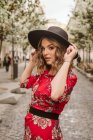 Sensual young woman in stylish dress and hat looking at camera while standing on aged pavement on city street — Stock Photo