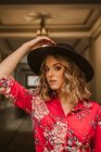 Стильна молода жінка в елегантній сукні і капелюсі дивиться на камеру, стоячи під лампою в проходженні старої будівлі — стокове фото