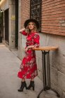 Mujer joven de moda en vestido elegante y sombrero mirando hacia otro lado mientras se apoya en la mesa redonda cerca del antiguo edificio en la calle de la ciudad - foto de stock