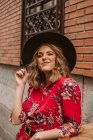 Joyeuse jeune femme en robe élégante et chapeau regardant la caméra près de vieux bâtiment sur la rue de la ville — Photo de stock