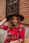 Attraente giovane donna in abito elegante e cappello guardando la fotocamera vicino vecchio edificio sulla strada della città — Foto stock