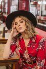 Sensuale giovane donna in elegante abito elegante e cappello seduto sul divano rosso vicino al tavolo e guardando la fotocamera nel ristorante — Foto stock