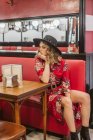 Jovem atraente em elegante vestido elegante e chapéu sentado no sofá vermelho perto da mesa no restaurante — Fotografia de Stock