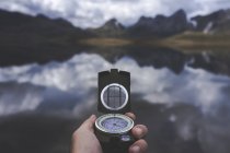 Mano di viaggiatore anonimo che tiene bussola contro il tranquillo lago di montagna in una giornata nuvolosa nella campagna spagnola — Foto stock