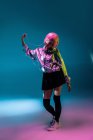 Jovem mulher asiática com corte de cabelo rosa elegante e jaqueta de prata brilhante em pé sobre fundo colorido e ouvir música — Fotografia de Stock