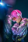 Fashionable attractive young Asian woman with pink hair in silver jacket listening music on dark background with reflection — Stock Photo