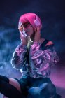Jovem mulher asiática elegante com cabelo roxo ouvindo música com fones de ouvido enquanto sentado no chão no fundo escuro neblina — Fotografia de Stock