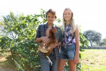 Teen Junge und Mädchen in karierten Hemden und Jeanshosen lächeln und streicheln Henne, während sie an einem sonnigen Tag auf einem Bauernhof neben grünen Büschen stehen — Stockfoto