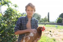 Jeune garçon et en chemise à carreaux et denim court poule caressant tout en se tenant près des buissons verts sur une journée ensoleillée à la ferme — Photo de stock