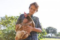 Jeune garçon et en chemise à carreaux et denim court poule caressant tout en se tenant près des buissons verts sur une journée ensoleillée à la ferme — Photo de stock