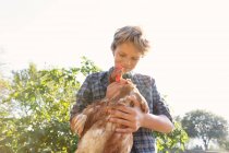 Teen boy and in checkered shirt and denim short petting hen while standing near green bushes on sunny day on farm — Stock Photo