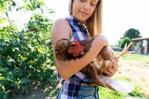 Chica adolescente y en camisa a cuadros y denim gallina de mascotas cortas mientras está de pie cerca de arbustos verdes en el día soleado en la granja - foto de stock